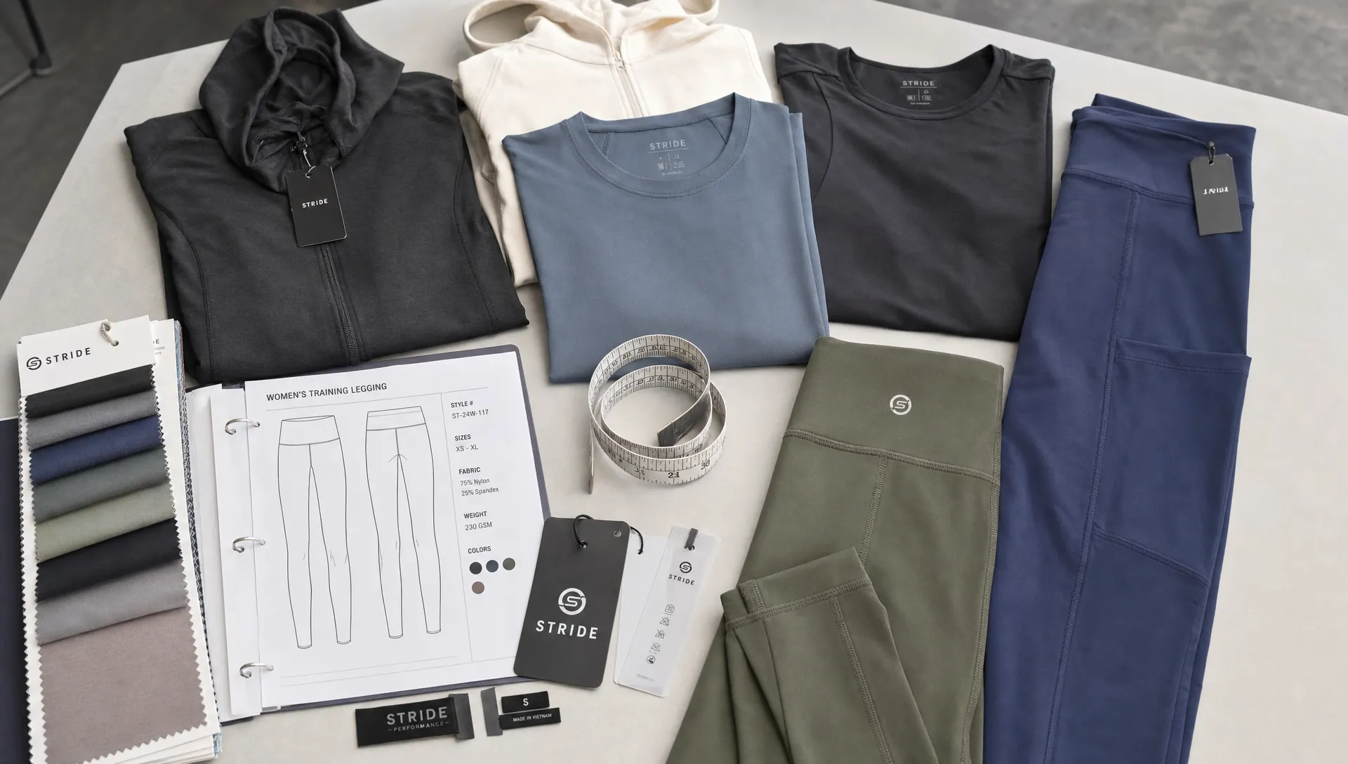 A collection of activewear samples arranged on a studio table, including leggings, training shirts, hoodies, fabric swatches, a measurement tape, and branded labels prepared for manufacturer review.